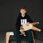 Junge sitzt auf Bank mit Holzspielzeug, lächelt, dunkler Hintergrund.