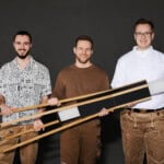 Drei Männer mit Holzschlitten vor grauem Hintergrund, lachend und in Freizeitkleidung.