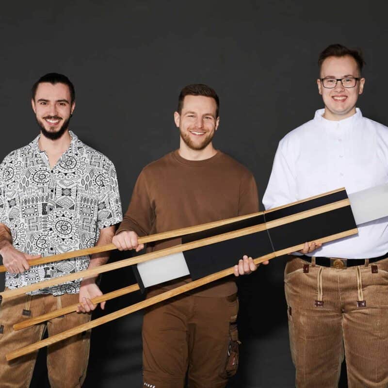 Drei Männer Mit Holzschlitten Vor Grauem Hintergrund, Lachend Und In Freizeitkleidung. Drei Männer Mit Holzschlitten Vor Grauem Hintergrund, Lachend Und In Freizeitkleidung.