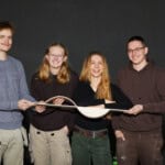 Gruppe junger Menschen hält ein Holzobjekt vor dunklem Hintergrund.