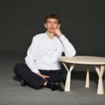 Junge sitzt entspannt neben einem runden Holztisch auf dunklem Hintergrund, zeigt Interesse an nachh.