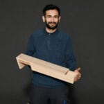Handwerker hält ein großes Holzstück vor schwarzem Hintergrund, Fokus auf Holz und Handarbeit.