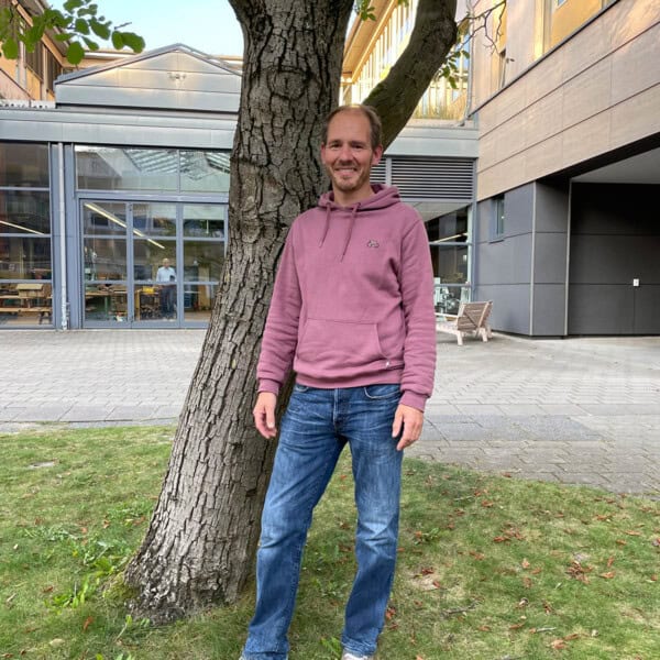 Mann In Rosa Hoodie Steht Unter Baum Vor Moderner Gebäudefassade, Natur Und Architektur Vereint.