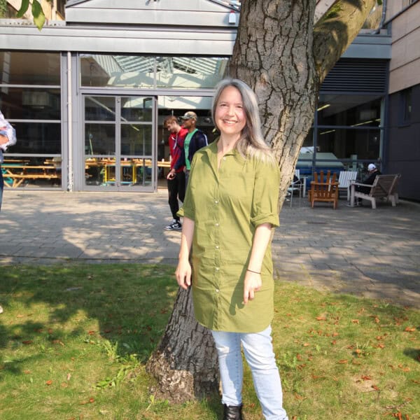 Fröhliche Frau Steht Lachend Am Baum Im Garten, Umgeben Von Natur Und Moderner Architektur.